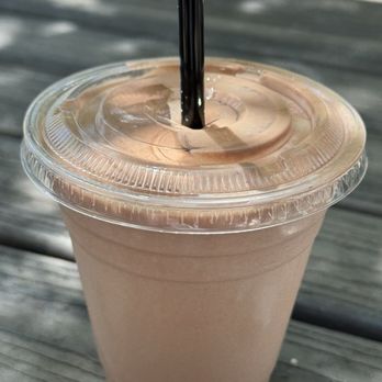Birdcall - All Natural Chicken - Chocolate Milkshake - A chocolate milkshake in a transparent plastic cup with a clear plastic lid and a black straw. The cup is on a weathered wooden surface, with sunlight and shadows visible in the background, indicating an outdoor setting. - Chicken Shop Near Me - Denver, Colorado