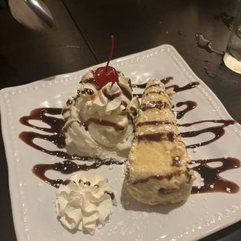 Shibui - Fried cheesecake with ice cream - A square white plate holds a slice of cheesecake drizzled with chocolate sauce, accompanied by a scoop of vanilla ice cream topped with whipped cream and a cherry. Additional whipped cream is placed on the plate, and the dessert is garnished with powdered sugar. The plate is set on a dark wooden table, with a glass of water and part of a person's arm visible in the background. - Japanese Near Me - Sunset, Florida