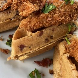 Two chicken and waffles with fried chicken, garnished with herbs, served on a white plate with some bacon bits and greens.