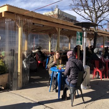 Le Paddock - Outdoor dining - An outdoor dining area with a wooden frame and transparent plastic walls, providing shelter for patrons. Several people are seated at tables, some engaged in conversation and others eating or drinking. The setting is on a sidewalk with parked cars along the street, and a heater is visible, indicating it may be a cooler day. The scene includes a yellow horse crossing sign and a green sign with a horse icon, suggesting a horse-themed or equestrian-related location. - French Near Me - Brooklyn, New York