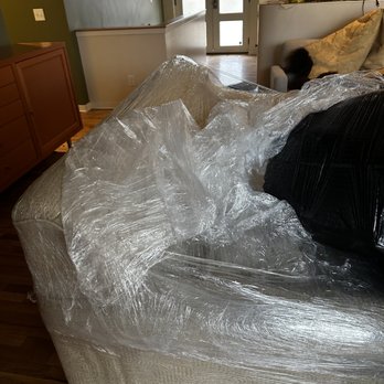 Fordson Movers - More plastic wrap - A beige upholstered sofa is wrapped in clear plastic in a living room, with a black cushion on it and a wooden floor. - Movers Near Me - Dearborn Heights, Michigan