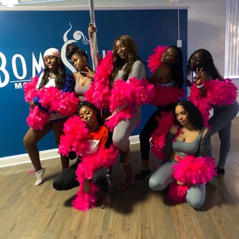Bombshell Movement Studio - Eight women pose together in a dance studio with a blue wall and a pole, wearing colorful clothing and pink feather boas. - Pole Dancing Classes Near Me - Chicago, Illinois