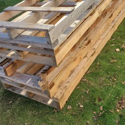 Heavy useless pallets - Multiple wooden pallets and planks are stacked outdoors on a grassy lawn, with a wooden fence in the background.