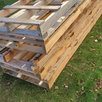 Junk King Rhode Island - Heavy useless pallets - Multiple wooden pallets and planks are stacked outdoors on a grassy lawn, with a wooden fence in the background. - Junk Removal & Hauling Near Me - Cranston, Rhode Island
