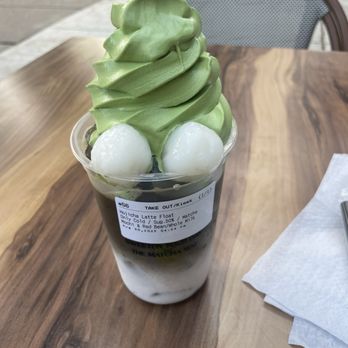 Kyo Matcha - A matcha latte float topped with green soft serve, mochi, and red bean in a clear cup, placed on a wooden table outdoors. - Desserts Near Me - Brookline, Massachusetts