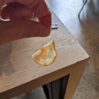 Vester - A HAIR! Cooked into the chip??! - A hand is holding a single potato chip by the edge, with the chip hanging downward. The chip is golden-brown and slightly curved. The background shows a wooden table with a metal clip placed on it, and part of a chair leg is visible in the upper right corner. The setting appears to be indoors, possibly in a casual dining or cafe environment. - Cafes Near Me - Cambridge, Massachusetts