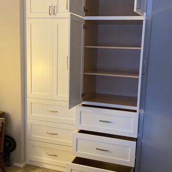 JC Custom Cabinets - Finished cabinet, doors and drawers opened - A white storage cabinet with two upper doors open, revealing three interior shelves. Below, there are three large drawers with metallic handles. The cabinet is positioned against a wall with beige carpet flooring, and part of another piece of furniture is visible on the left side of the image. - Cabinetry Near Me