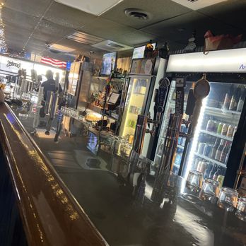 Bob's Bar - The image shows the interior of a bar with a polished wooden counter, beer taps, and refrigerators filled with beverages. The ceiling has acoustic tiles and ventilation systems. In the background, patrons are seen near the entrance, which is bright with natural light. The bar area includes various bottles and bar equipment, with a television mounted on the wall. - Bars Near Me - Columbus, Ohio
