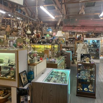 Bougie Bazaar - Unique collection - The image shows the interior of a vintage thrift store filled with collectibles, jewelry, art, and home decor items. Display cases with glass tops contain various small items, while shelves and hanging racks hold decorative objects, framed pictures, and accessories. The store has a rustic wooden ceiling with exposed beams and bright fluorescent lighting. The space appears densely packed with merchandise, creating an eclectic and nostalgic atmosphere. - Antiques Near Me - Pasadena, California