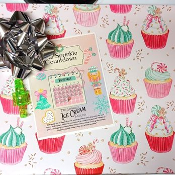 Mrs. Sprinkles Ice Cream - Ice Cream Advent Calendar packaging. So cute! - A gift box wrapped in cupcake-patterned paper features a silver bow, a green plastic toy, and a holiday-themed sticker sheet with a calendar and decorations. - Ice Cream & Frozen Yogurt Near Me - Albuquerque, New Mexico