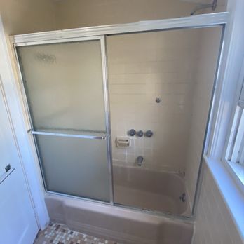 Sun Shore Construction - Old bathroom - A bathroom shower features frosted glass sliding doors, beige tiled walls, and a small bathtub. The showerhead is mounted on the wall, with control knobs below. - General Contractors Near Me - Needham, Massachusetts