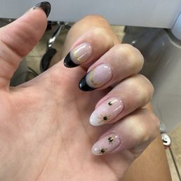 SNS with design done by Trinity! - Close-up of a hand with decorated nails featuring black tips, gold lines, and bee motifs, likely at a nail salon.
