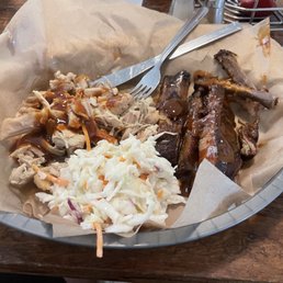 Pulled Chicken - A tray lined with parchment paper holds a barbecue meal including pulled chicken with barbecue sauce, pork ribs with sauce, and a serving of coleslaw with shredded cabbage and carrots. A fork and a knife are placed on the tray, and the tray is on a wooden table. The setting suggests a casual dining environment at a barbecue restaurant.