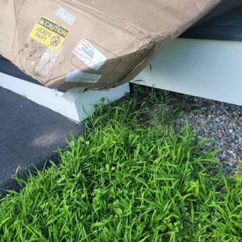 McGee Toyota of Epping - Another smashed portion of my product - A large cardboard box with visible damage and caution labels is positioned on a white platform. The box appears to be heavy and is partially crushed. Surrounding the platform, there is overgrown green grass and small gravel, with a section of asphalt pavement nearby. The scene suggests outdoor storage or delivery packaging that has been left outside. - Car Dealers Near Me - Epping, New Hampshire