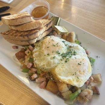 Daily Jam - Breakfast & Brunch Restaurant - The Denver Sauté - A breakfast plate includes two fried eggs garnished with chopped herbs, placed over a mixture of diced vegetables and meat. Accompanying the eggs are three slices of toasted bread with grill marks, and a small container of syrup. The dish is served on a rectangular white plate, with a glass of water visible in the background on a wooden table. - New American Near Me - Tempe, Arizona