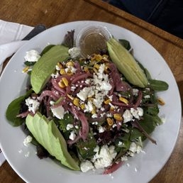 Avocado Salad - A mixed green salad with avocado slices, pickled red onions, corn, and crumbled cheese, served with a small container of dressing on a white plate.