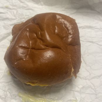 Dengeos - Fish - The image features a single burger bun with a shiny, brown top, resting on crinkled white paper. The bun appears soft and slightly uneven in shape. There is a small amount of melted cheese visible at the bottom edge of the bun, with some cheese residue on the paper. The background is plain and neutral, emphasizing the bun as the main subject. - Greek Near Me - Buffalo Grove, Illinois