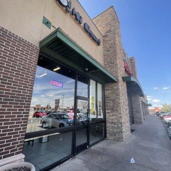 Hi BoBa - The image depicts the exterior of Hi BoBa, a boba tea shop located in Albuquerque, NM. The storefront features a glass door with promotional posters and an illuminated 'OPEN' sign. The building has a combination of brick and stone exterior walls, with a sidewalk in front and parked cars visible across the street. The sky is clear and blue, indicating a sunny day. - Bubble Tea Near Me - Albuquerque, New Mexico