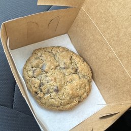 Oatmeal Chocolate Chip Cookie - A sizable oatmeal chocolate chip cookie is placed in a brown bakery box with a piece of parchment paper underneath.