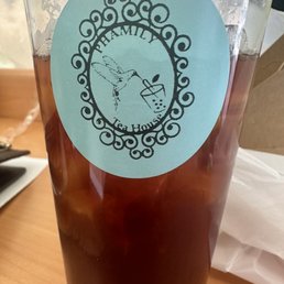 Winter Melon Tea - A tall, transparent glass filled with iced tea and ice cubes is placed on a wooden table. The glass has a blue oval label with the logo of Phamily Tea House, featuring a hummingbird drinking from a bubble tea cup, surrounded by decorative swirls. The setting appears to be indoors, with a napkin and part of a plate visible nearby.