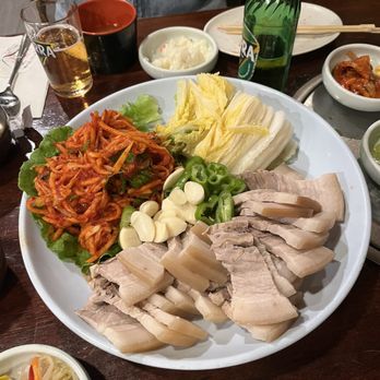 Yechon - bossam (pork belly) - The image shows a large round white plate with Korean cuisine, including boiled pork belly slices, kimchi, lettuce, garlic cloves, and sliced green peppers. The table also has side dishes, a glass of beer, a green bottle, and other small bowls with various Korean foods. The setting appears to be a Korean restaurant with a wooden table and chopsticks. - Korean Near Me - Annandale, Virginia