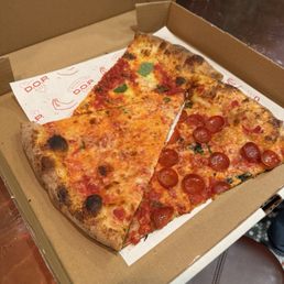 Pizza slices - A cardboard pizza box contains two slices of pizza, one with pepperoni and the other plain cheese. The slices have a thin, crispy crust with melted cheese and toppings. The box is placed on a table with a dark surface, and part of a red container is visible in the background.