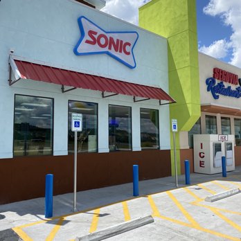 TA Express - Sonic inside - The image shows the exterior of a Sonic fast-food restaurant with a large sign displaying the Sonic logo. The building has a white facade with a red awning over the windows. There are two reserved parking signs indicating accessible parking spaces, marked with yellow lines and blue posts. An ice vending machine is visible near the entrance. The sky is partly cloudy with blue skies and white clouds, and the overall scene suggests a typical drive-in restaurant setting. - Gas Stations Near Me - Junction, Texas