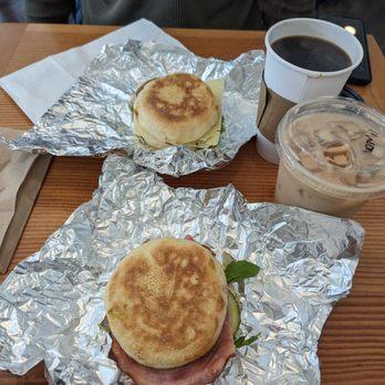 Vinal Bakery - Two breakfast sandwiches wrapped in aluminum foil are on a wooden table, accompanied by a cup of coffee and an iced beverage. - Cafes Near Me - Somerville, Massachusetts