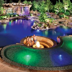 Blue Haven Pools & Spas - Lagoon-style concrete pool with integrated landscape pockets, sunken conversation area with fire pit, and multicolor LED lighting