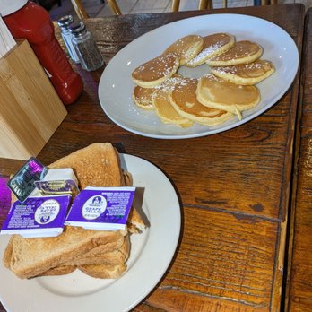 Cantina 35  - Silver dollar pancakes and whole wheat toast - The image shows a wooden table with two plates of breakfast food. One plate contains several pancakes dusted with powdered sugar, while the other plate has toasted bread slices with packets of grape jelly on top. Condiment shakers and a ketchup bottle are also visible on the table, along with a wooden napkin holder. The background includes chairs and part of a restaurant interior. - Mexican Near Me - Queens, New York