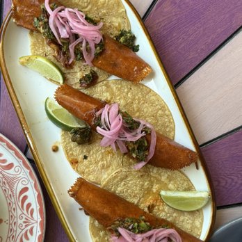 Mariscos San Pedro - Shrimp Gobernador - A white oval plate holds three fish tacos, each with a fried fish fillet wrapped in a corn tortilla. The tacos are garnished with pickled red onions and chopped green herbs. Two lime wedges are placed on the plate for added flavor. The plate is set on a purple and beige wooden table, with part of a decorative plate visible on the side. - Mexican Near Me - Chicago, Illinois