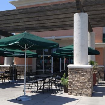 Starbucks - The image shows an outdoor seating area of a Starbucks coffee shop with several black metal tables and chairs. Green umbrellas with the Starbucks logo provide shade. The background includes the building's exterior with stone accents and white columns, and a large window reflecting the interior. The setting appears to be a sunny day with clear skies. - Coffee & Tea Near Me - Temecula, California