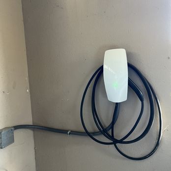 S&E Electric - A Tesla EV charger is mounted on a wall, with cables coiled around the device, in an indoor setting with a power outlet nearby. - Electricians Near Me - Auburn, California