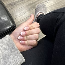 French round SNS manicure  (no tips or anything) - A person with a French manicure on their nails is seated, with their hand resting on their lap, wearing a beige sweater and black pants.