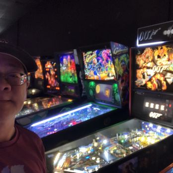 Electric Bat Arcade - Pinball machines - A man with glasses and a cap is taking a selfie at an arcade featuring pinball machines with colorful artwork and lighting. - Arcades Near Me - Tempe, Arizona