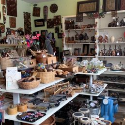Different things like Statues, salt lamps,etc. - The shop interior showcases a variety of handmade products including jewelry, wooden sculptures, masks, and decorative artifacts. Items are displayed on white tables and shelves, with some hanging on the walls. The shop has a ceiling with fluorescent lighting and a light green wall background, emphasizing the diverse collection of artisanal goods.