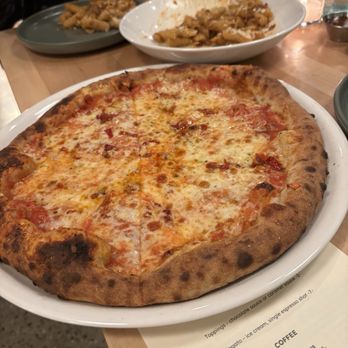 Myrth - Spicy cheese pizza - A cheese pizza with a golden, slightly charred crust and melted cheese on a white plate, with other dishes and drinks visible on the table. - Pizza Near Me - Ada, Michigan