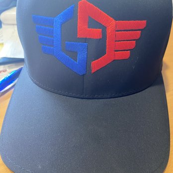 Image On - A black baseball cap features embroidered logos in blue and red, with winged designs on the front panel, placed on a wooden surface. - Screen Printing/T-Shirt Printing Near Me - Santee, California