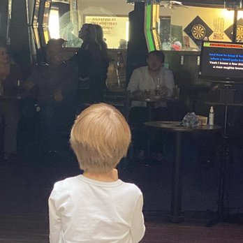 The Embers - A child with blonde hair, wearing a white shirt, faces a karaoke stage in a bar, with adults and dartboards visible in the background. - Salad Near Me - Eugene, Oregon