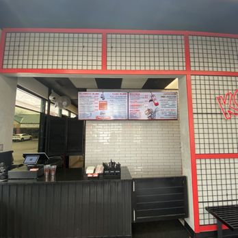 SubKO - Walk up counter to order - The image shows a restaurant counter with two digital menu screens displaying food options. The counter has a cash register, utensils, and a tip jar. The interior features white brick walls, black accents, and a grid-patterned wall with red framing. Large windows on the left side allow natural light into the space. - Sandwiches Near Me - Lisle, Illinois