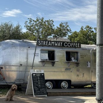 Streamway Coffee - A silver Airstream trailer is set up as a coffee truck with a sign that reads 'Streamway Coffee' on top. The trailer has two service windows, one labeled 'Order Here' and the other 'Pick Up Here.' A menu board is placed outside on a stand, listing various coffee options. A dog is sitting on the ground nearby, and a person is partially visible on the left side of the image. The background includes trees and a partly cloudy sky, indicating an outdoor setting near Mueller Lake Park in Austin, TX. - Coffee & Tea Near Me - Austin, Texas