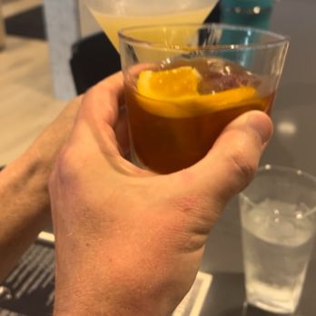The Kitchen - Cheers! - A person is holding two cocktails, one with a lemon slice and dark liquid, and another with a yellowish drink, in a bar or restaurant. The background shows a modern interior with tables, chairs, and large windows. A glass of water is on the table, and the person’s hand is visible. The setting appears casual and social. - Bars Near Me - Carmel, Indiana