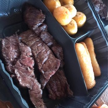 Vacunos - Churrasco - Papas Criollas - Yuca Fries - A black plastic tray with three compartments displays grilled steak on the left, bread rolls in the upper right, and fried cheese sticks in the lower right. The steak appears cooked with a seared exterior, and the bread rolls are golden-brown. The fried cheese sticks are elongated and crispy. The tray is placed on a wooden surface, and part of a drink container is visible at the bottom right corner. - Steakhouses Near Me - Riverview, Florida