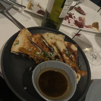 Root & Flower - Quesadilla - A plate of quesadilla with melted cheese, vegetables, and sauce is placed on a black serving dish. A small bowl of dark sauce accompanies the quesadilla. The table has wine bottles, plates with leftover food, and utensils, indicating a dining setting at a restaurant. - Wine Bars Near Me - Vail, Colorado