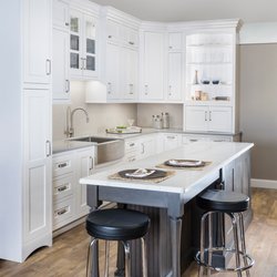 Kitchen Views - Kitchen showroom display featuring white cabinetry, a stainless farmhouse sink, an open display hutch with glassware, quartz ...
