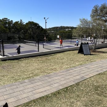 Frontyard Brewing - Pickle Ball courts - The image shows several people playing pickleball on outdoor courts enclosed by a chain-link fence. The courts are situated in a park-like setting with trees and a clear blue sky overhead. A signboard is placed on the grass near the courts, and a paved walkway runs alongside the area. The scene includes players of different ages actively participating in the game, with some holding paddles and a ball in play. - Breweries Near Me - Spicewood, Texas