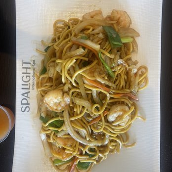 Spa Light - A rectangular white plate contains stir-fried noodles with shrimp, sliced onions, carrots, and green onions, coated in a savory sauce. The plate is placed on a dark surface, with part of a drink visible on the side. The dish appears to be a Chinese or Asian-inspired noodle entrée served at Spa Light, a Korean-inspired spa offering various relaxation amenities. - Massage Near Me - Lincoln, Rhode Island