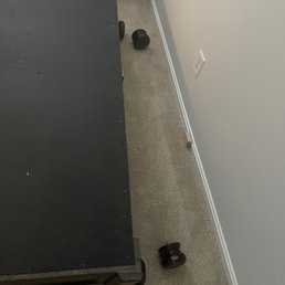 Like how you break the dresser in the same spot on both ends and don't take any responsibility?! - The image shows a black furniture item with wheels situated against a wall with a white electrical outlet and a beige carpeted floor. Several dark wooden knobs are scattered on the carpet near the furniture, with some close to the wall and others near the furniture's wheels. A cardboard box is partially visible in the background, indicating an indoor setting, possibly a storage or moving area.