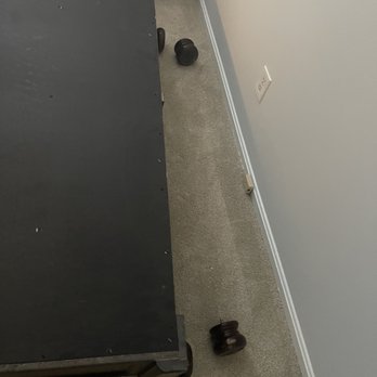 ZMax Movers - Like how you break the dresser in the same spot on both ends and don't take any responsibility?! - The image shows a black furniture item with wheels situated against a wall with a white electrical outlet and a beige carpeted floor. Several dark wooden knobs are scattered on the carpet near the furniture, with some close to the wall and others near the furniture's wheels. A cardboard box is partially visible in the background, indicating an indoor setting, possibly a storage or moving area. - Movers Near Me - Fairfax, Virginia