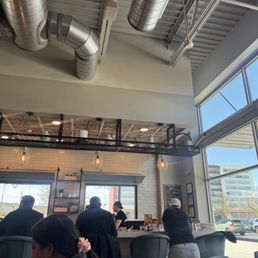 Inside - Inside a cafe, customers sit at a counter with a barista preparing drinks. The cafe features large windows, industrial ceiling pipes, and modern lighting.